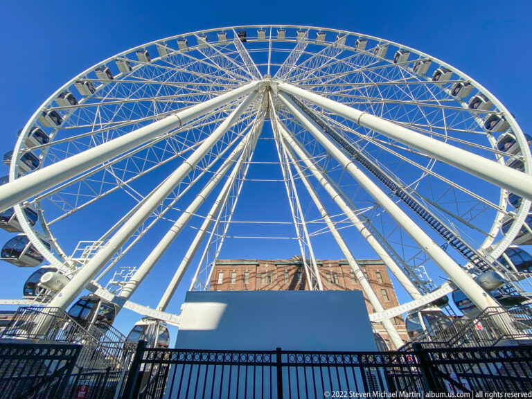 White Ferris wheel spokes converge above a brick building and fenced entrance against blue sky.