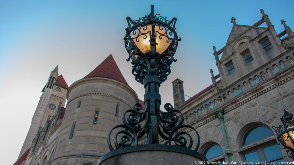 An ornate wrought-iron lantern stands before a stone turret with a red roof and arched windows.
