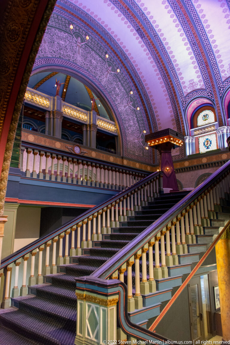A broad ornate staircase ascends beneath a patterned vaulted ceiling toward stained-glass windows and a balcony.