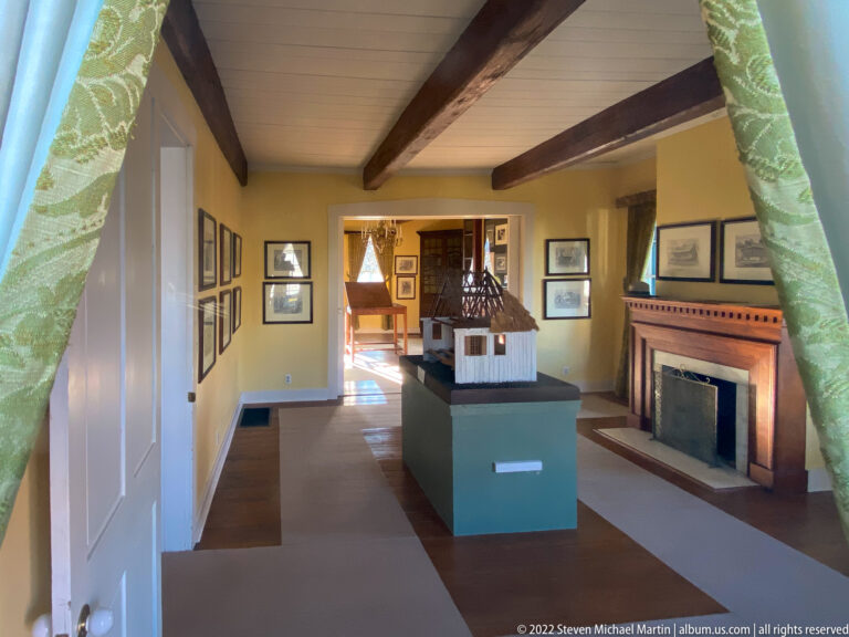 A building model stands on a pedestal in a beamed room with a fireplace and framed pictures.