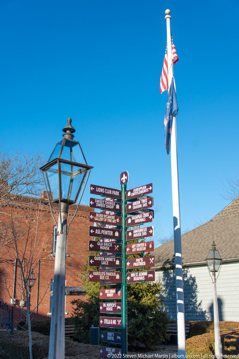 Directional signs point to local destinations beneath two flags beside a street lantern.