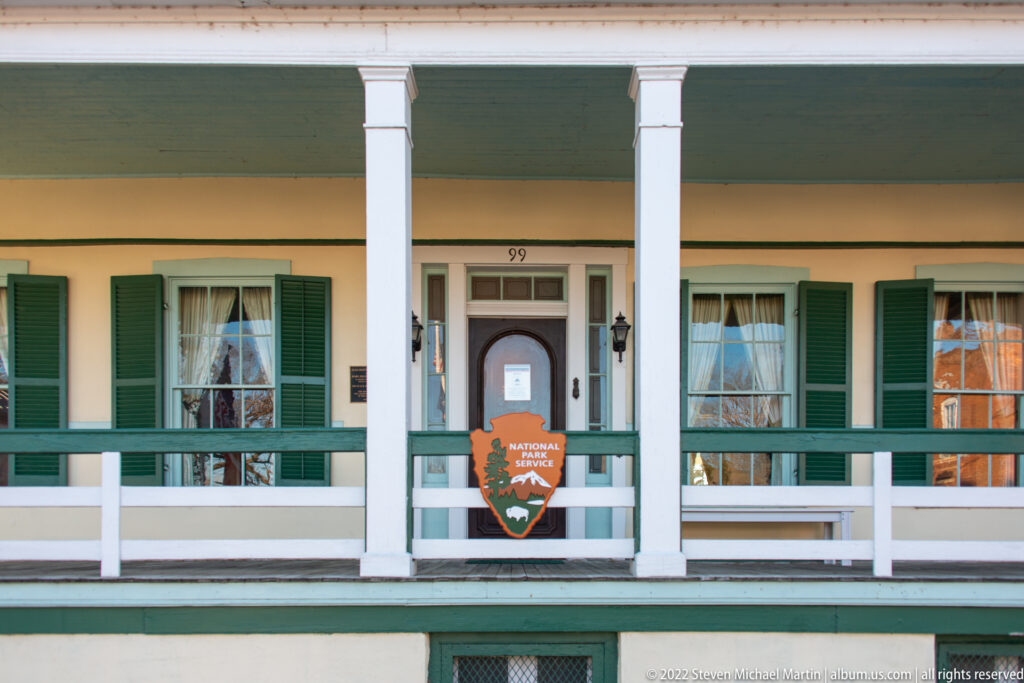 A clapboard house has green shutters, a long upper gallery, and an NPS emblem on its railing.