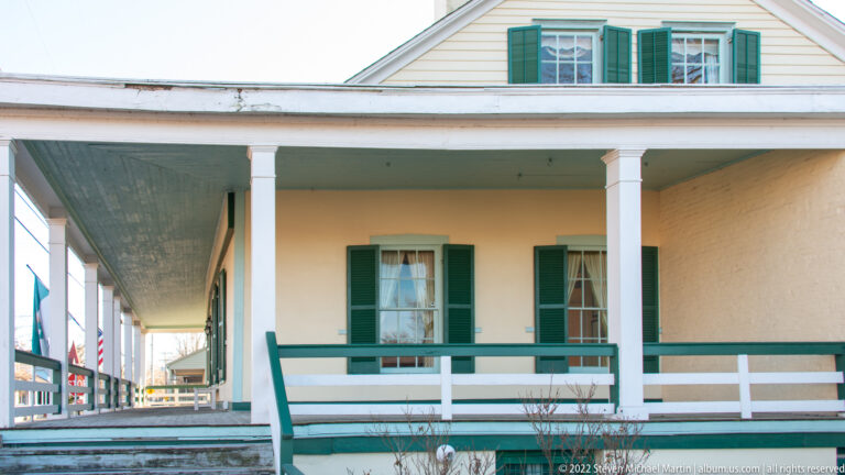A covered gallery runs along a yellow house with green shutters and white railings.