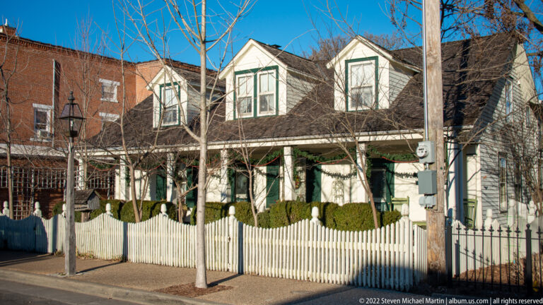 A white two-story house with projecting gables and a porch stands behind a picket fence.
