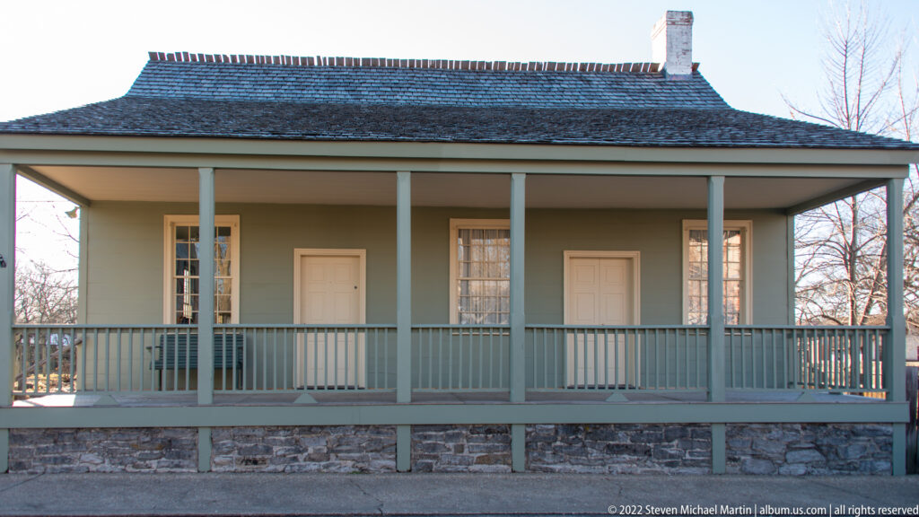 A low house has a long post-supported gallery, two doors, tall windows, and a stone foundation.