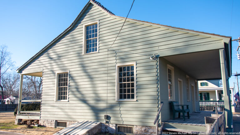A pale clapboard gable end with several windows joins a deep side gallery.