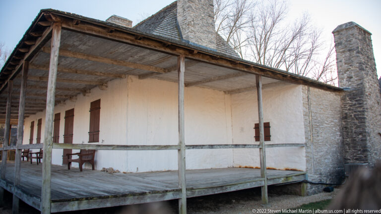 A weathered gallery wraps around a pale house beside a tall stone chimney.