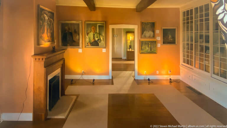 Framed portraits hang above a fireplace in an ochre room with a tall multi-pane window.
