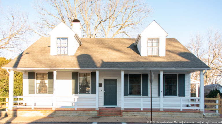 A clapboard house has two roof dormers, a central door, and a full-width porch.