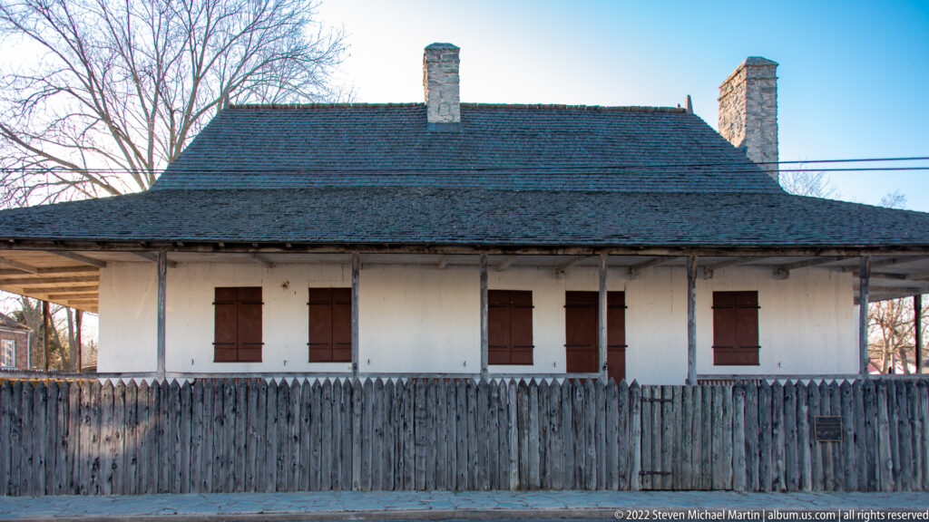 A pale one-story house with red shutters stands behind a tall wooden fence.