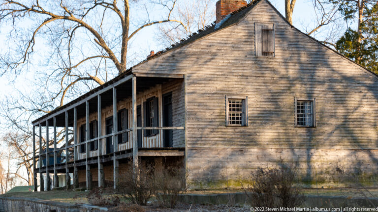 A weathered timber-sided house has a deep gallery, gabled end, and raised stone base.