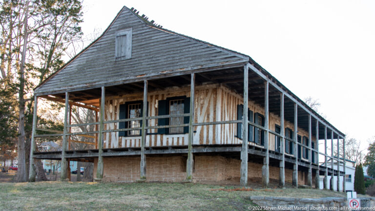 A raised timber house has exposed framing, a broad roof, and a long upper gallery.