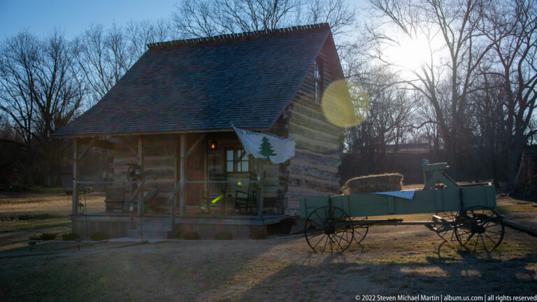 A wooden wagon stands beside a timber house in low winter sunlight.