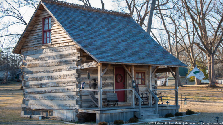 A log house with a steep shingled roof has a small porch, red door, and flag beside the entrance.
