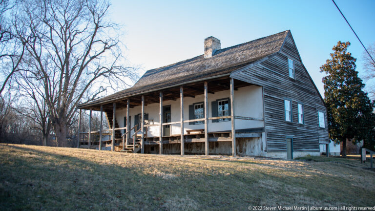 A two-story timber house with a steep roof and wraparound gallery stands on a stone base.