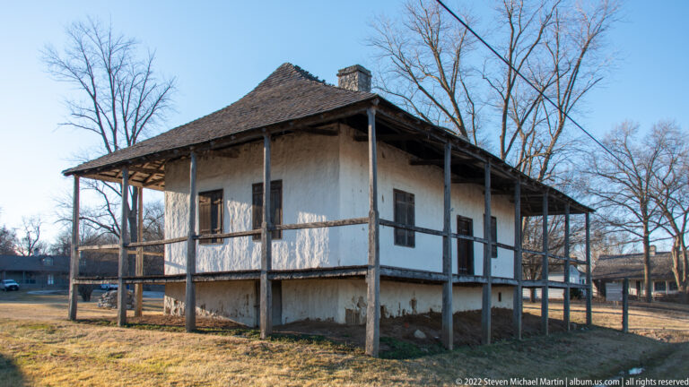 A pale raised house has a deep post-supported gallery beneath a broad projecting roof.