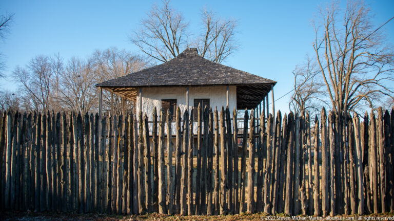 A tall pointed wooden fence partly hides a small raised house and covered porch.