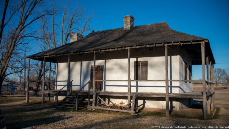 A pale timber house has a raised gallery, short steps, multi-pane windows, and a broad roof.