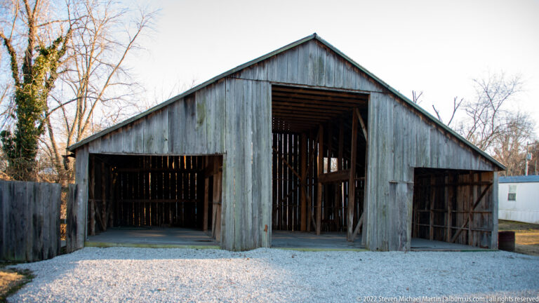 A weathered timber shed with three open bays faces a gravel forecourt among leafless trees.