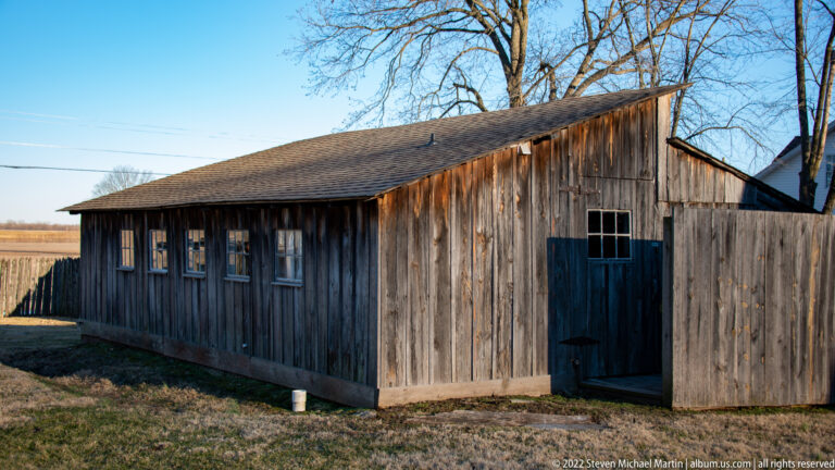 A long weathered wooden outbuilding has small windows and a dark door beneath a sloping roof.