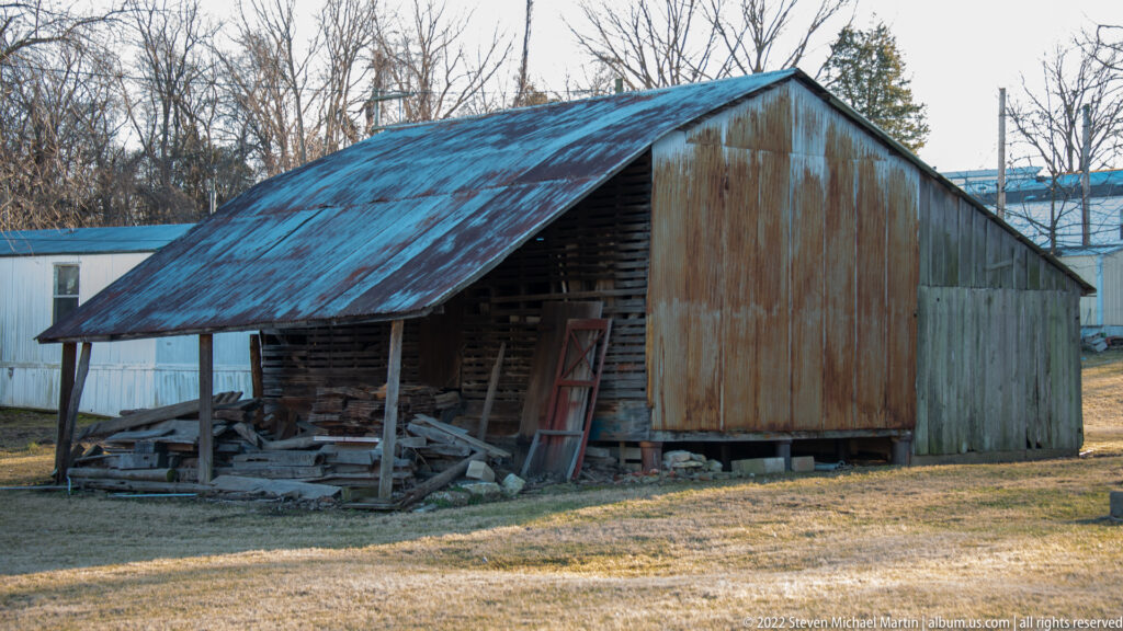 A rusted metal roof shelters stacked lumber beside the open bay of a weathered timber shed.