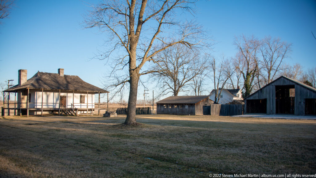 A raised house and several wooden outbuildings stand across an open winter yard beneath a leafless tree.