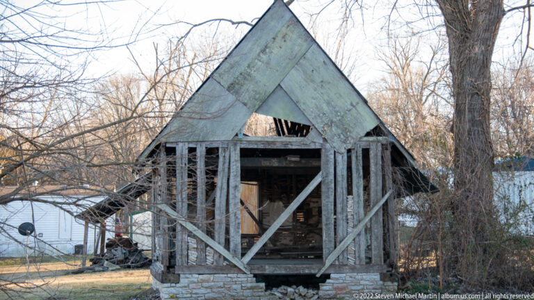 Exposed posts and diagonal braces form the gable end of a small timber building on a stone base.