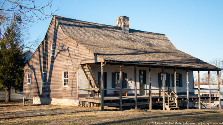 A long wooden house has a deep post-supported porch, steps, and a broad shingled roof.