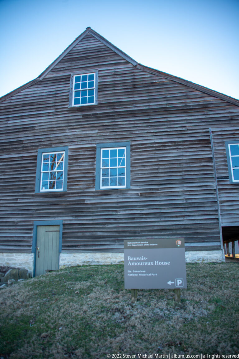 The dark, elongated Bauvais-Amoureux House stands behind a small identification sign on a grassy slope.