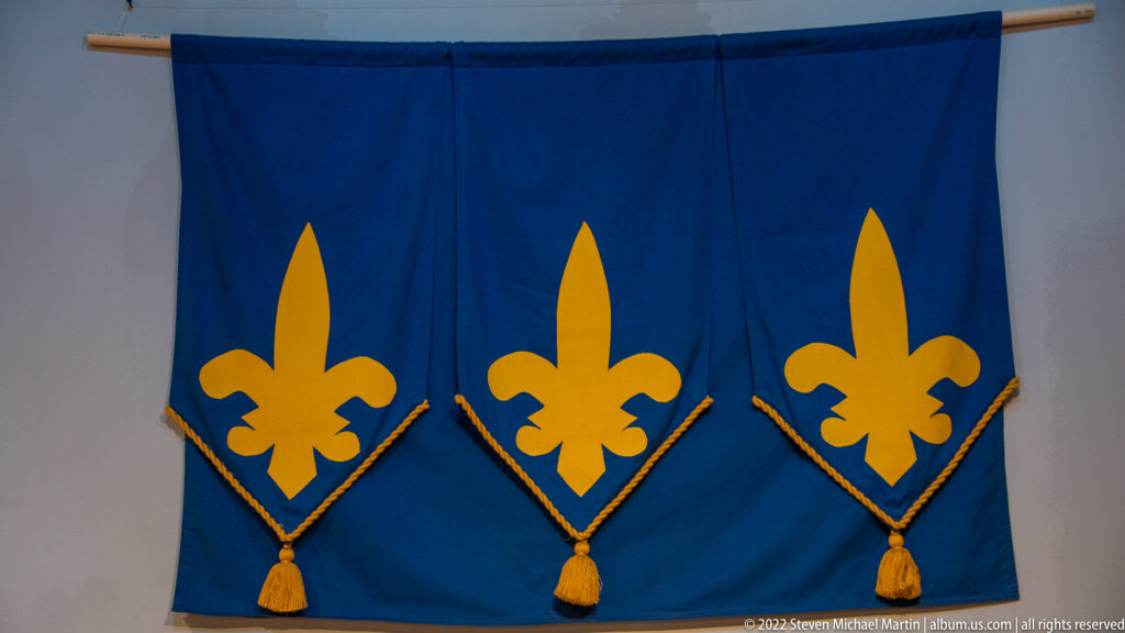 Three gold fleur-de-lis emblems decorate a blue banner with pointed borders and tassels.