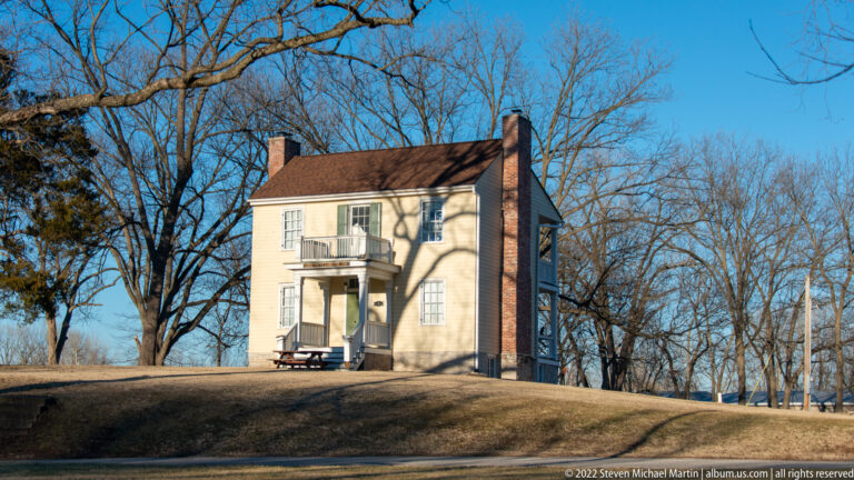 A white two-story house with a central balcony and brick chimney stands above a road on a grassy rise.