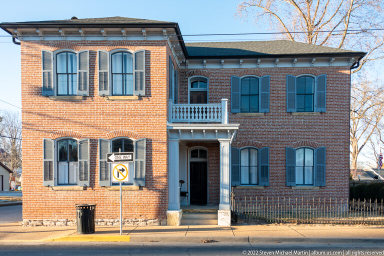 A two-story brick house has blue shutters, a central balcony, and a recessed doorway facing the street.