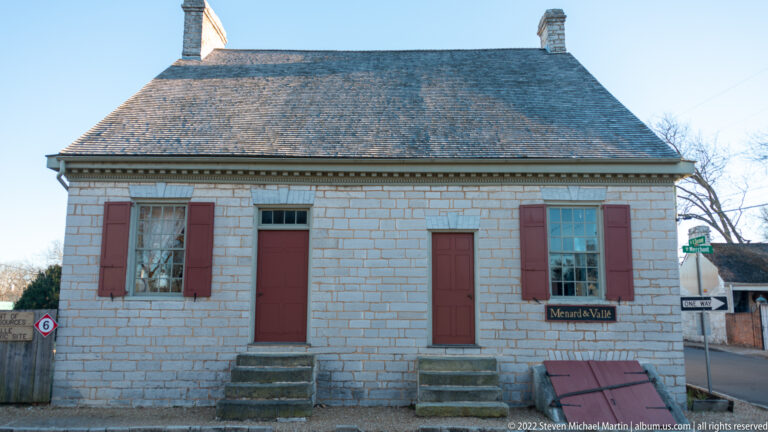A pale stone house has red shutters and a central red door beneath a steep roof.