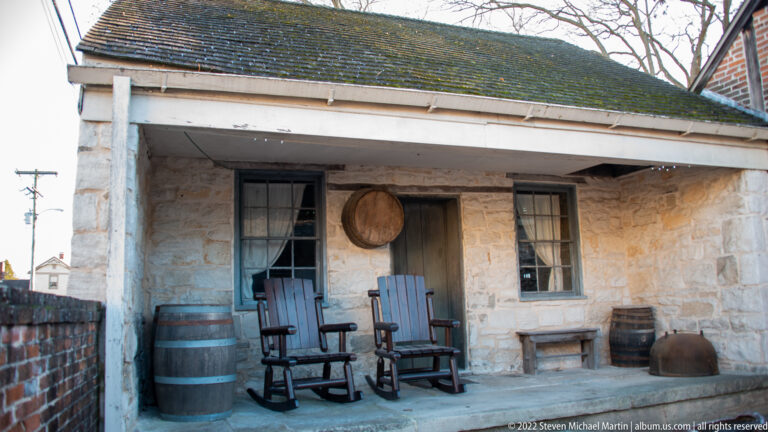 Two rocking chairs and wooden barrels sit on a covered porch against a rough stone wall.