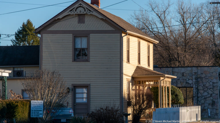 A narrow two-story clapboard house stands beside a stone wall with a car parked near its porch.