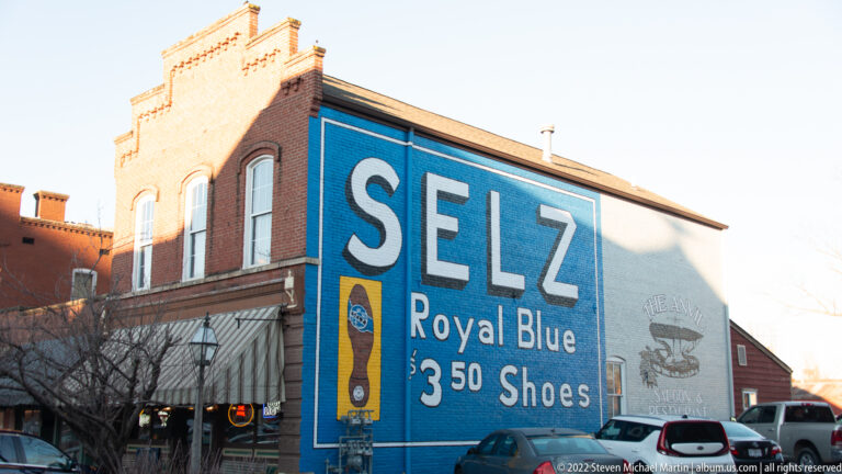 A large blue wall advertisement reads SELZ Royal Blue $3.50 Shoes on a brick storefront.