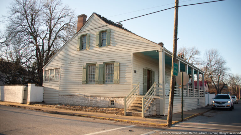 A white two-story house with pale green shutters has an extended covered porch along the street.