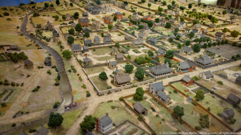 An aerial view of a scale model shows houses, gardens, roads, trees, and a stream.