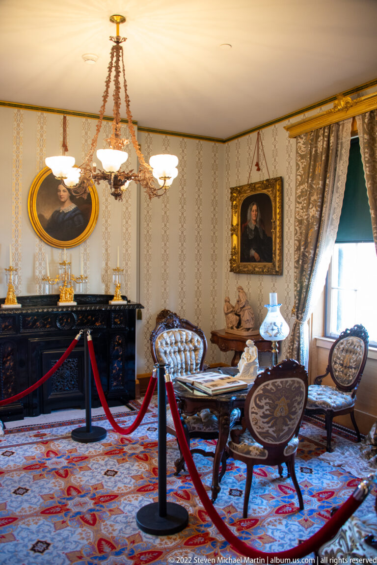 A formal parlor contains a fireplace, portraits, chandelier, patterned rug, and carved chairs.