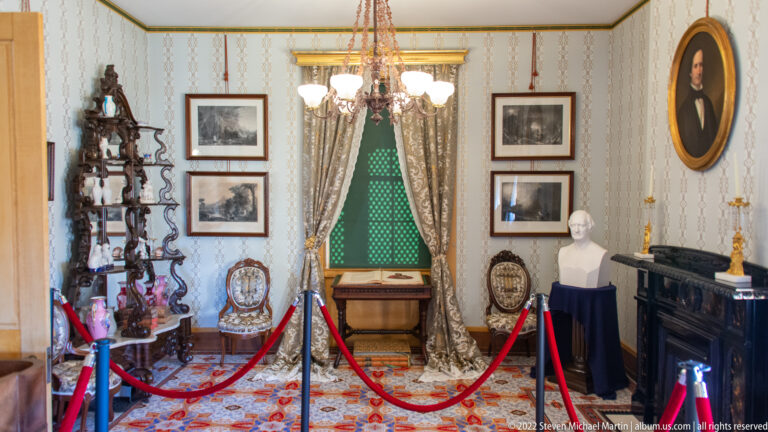 A historic room displays portraits, a bust, carved shelving, and a curtained window behind red ropes.