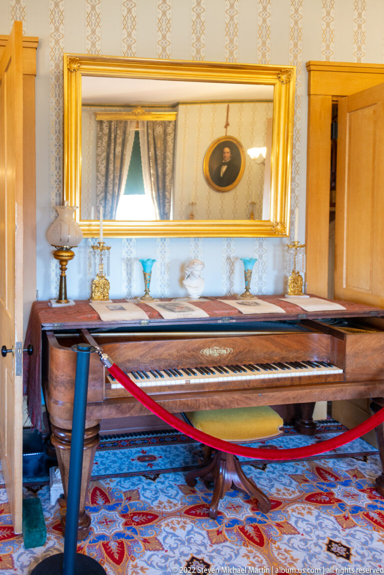 A polished grand piano stands beneath a gilt mirror reflecting a window and portrait.