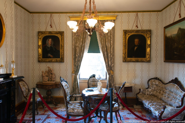Patterned chairs and a settee face a window beneath portraits in a furnished parlor.
