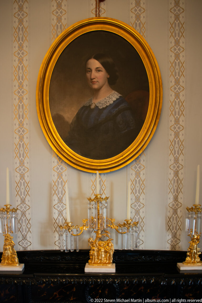 An oval portrait of a woman hangs above a black mantel with gilded candlesticks.