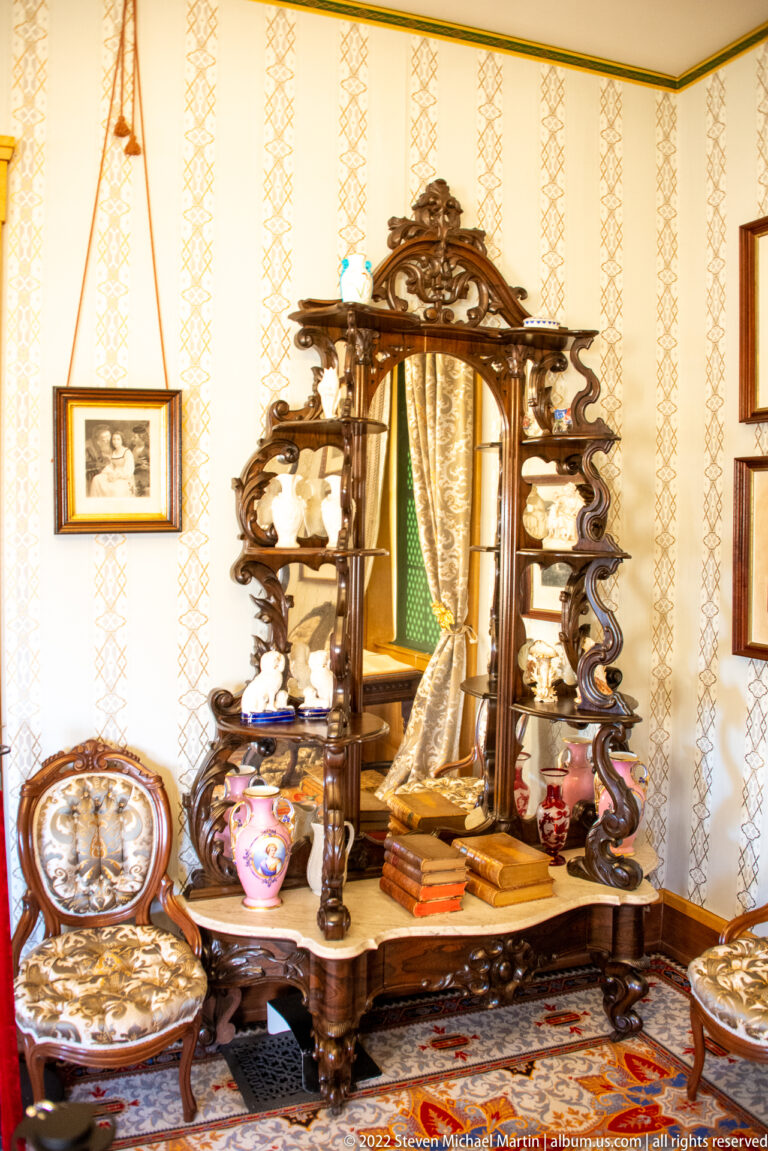 A carved mirrored cabinet stands above books, ceramic vessels, and decorative objects in a parlor.