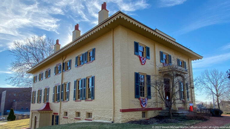 An angled view shows two brick facades with dark shutters, red trim, and small flags.