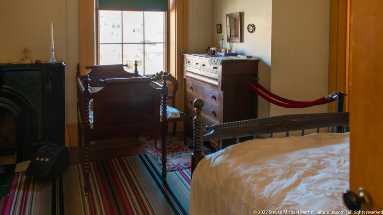 A bedroom contains a white bed, writing desk, chair, dresser, and striped rug.