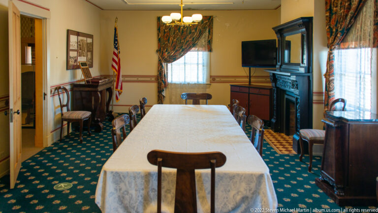 A long dining table fills a room with wooden chairs, curtained windows, a fireplace, and an American flag.