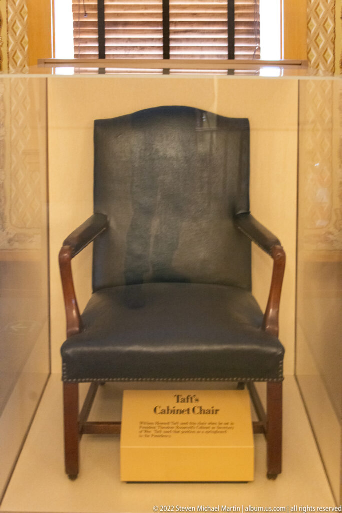 A dark upholstered armchair with curved wooden arms is displayed behind glass above its identification label.