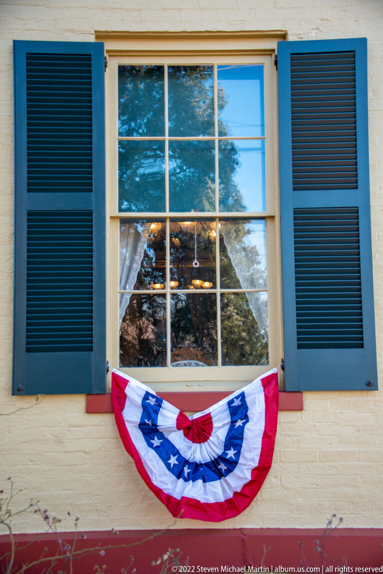Red, white, and blue bunting hangs beneath a window with blue shutters on a pale clapboard wall.