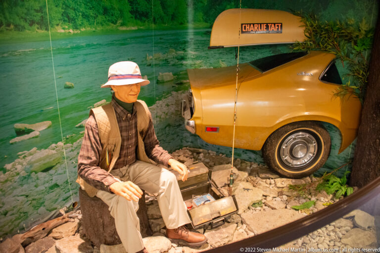 A seated mannequin in a hat and vest rests beside a tan car labeled CHARLIE TAFT in a staged display.